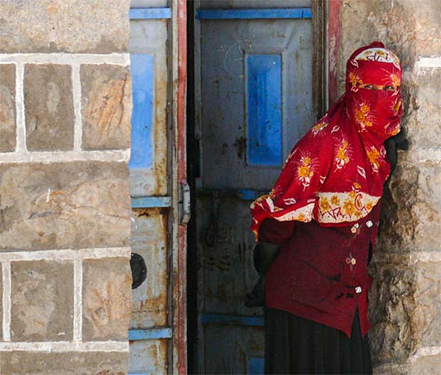 Yemeni culture: Village woman in a colorful red print headscarf is leaning stone wall in front of a blue door by Maarten de Wolf.
