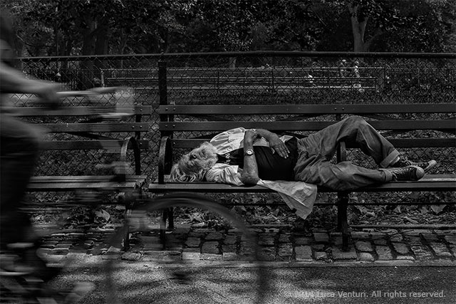Black and white image of a vagrant sleeping on a park bench in the gardens of the square in New York City by Luca Venturi.