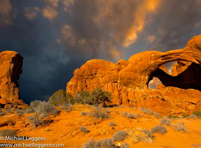 Storm clouds appearing over orange colored Double Arch rock formation at sunset at Arches National Park in Utah by Michael Leggero.
