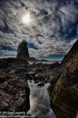 Dark stormy clouds and some sun over rock formations along shoreline in the Olympic Peninsula in Washington by Michael Leggero.