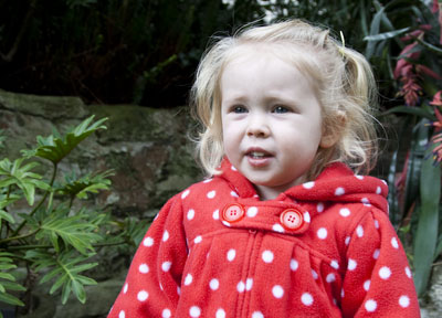 Close-up photo portrait of a toddler in a red and white polka dot top - outside in front of plants by Cathy Topping.