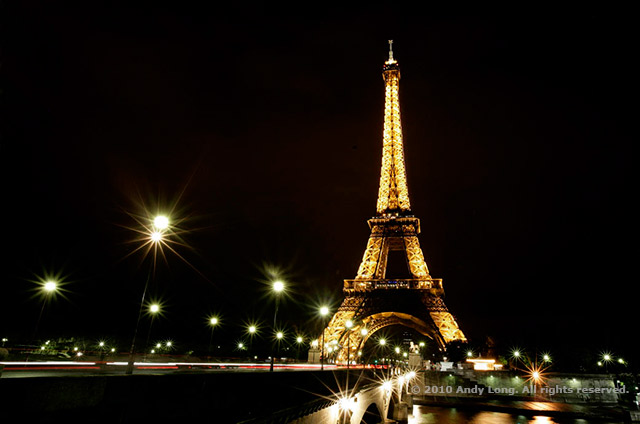 Image of the Eiffel Tower lit at night with leading lines of light posts and the lights from moving cars by Andy Long.