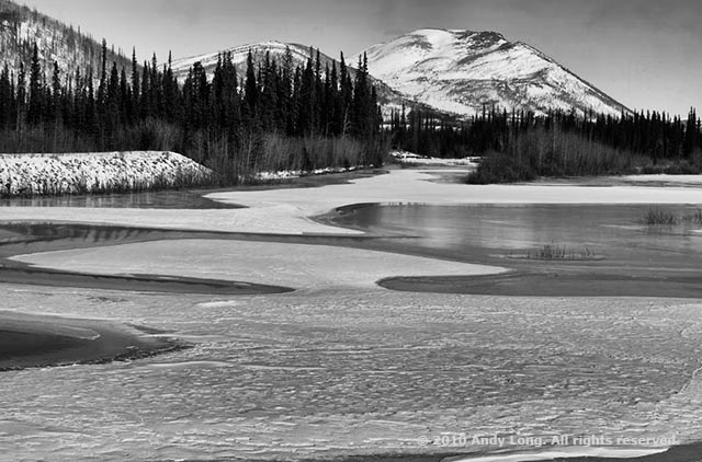 Black and white image showing curves in an icy pond that lead the eye to a mountain by Andy Long.