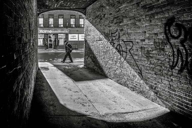 Black and white image created from inside a brick alley with a man walking past and harware store sign in the back by Randall Romano.