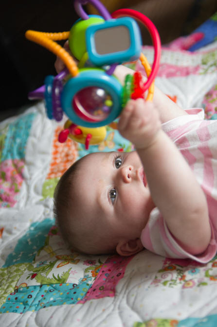 While laying on a quilt, Baby A plays with a colorful toy by Elizabeth Powis Fulks.